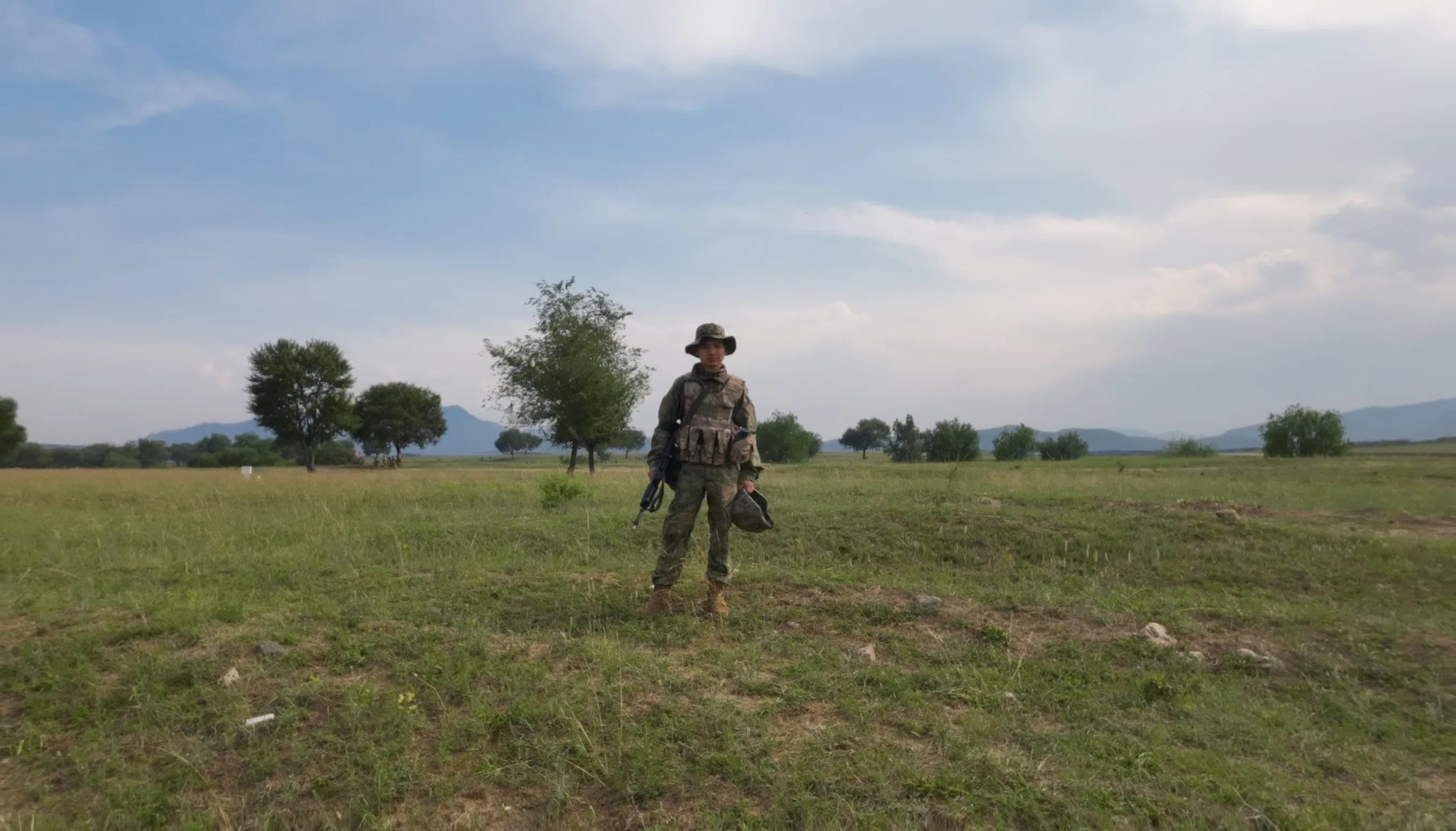 A person in camouflage gear standing on open grassland, trees and hills beyond